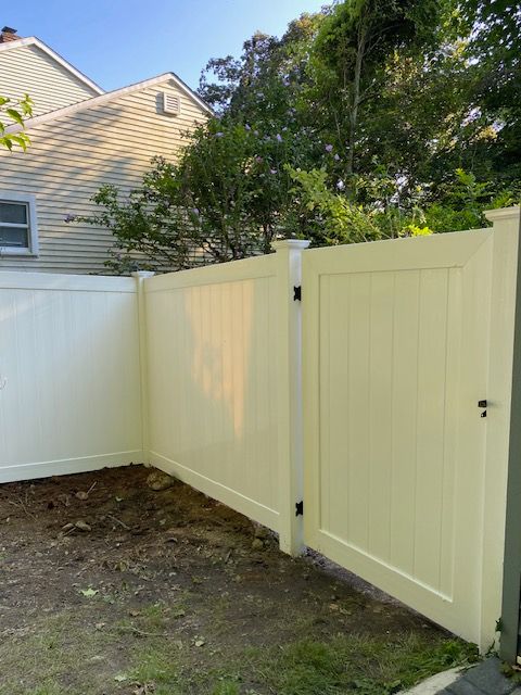 Cream-colored wooden gate beside a white fence in a backyard, with a house and trees in the background