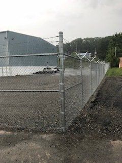 Chain-link fence around a gravel lot beside a large gray industrial building.