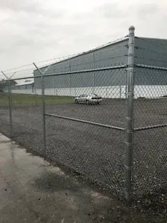 Chain-link fence beside a wet parking lot with a white car and a large gray industrial building.