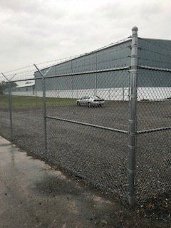 Chain-link fence beside a wet road with a white car parked near a large gray industrial building.