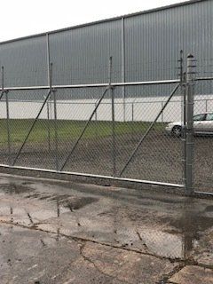 Chain-link fence and industrial building on a rainy day, with a wet pavement and parked car behind the fence.