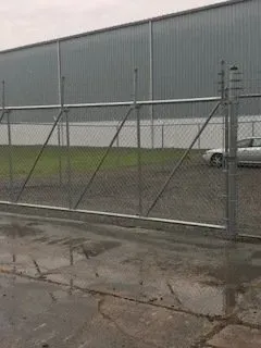 Chain-link fence in front of a gray warehouse on a wet, cracked asphalt lot, with a car at right.