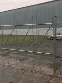 Chain-link fence beside a gray warehouse on a wet, rainy lot, with a car parked behind it.