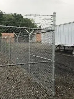 Chain-link fence topped with barbed wire beside a gravel yard and white trailer.