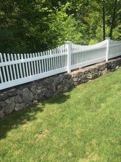 White picket fence atop a stone retaining wall along a green grassy slope beside trees.