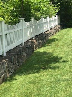 White scalloped fence atop a stone retaining wall beside a green lawn and trees