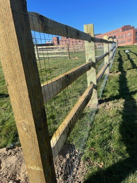 Wooden fence with wire mesh along a grassy path beside brick buildings on a sunny day