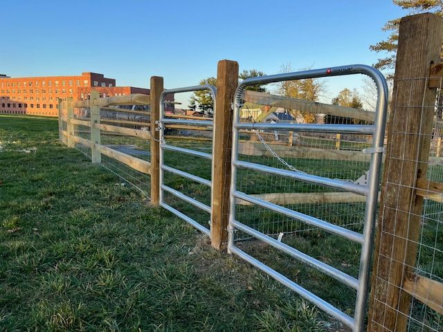 Metal farm gate and wooden fence along a grassy field at sunset, with a brick building in the background
