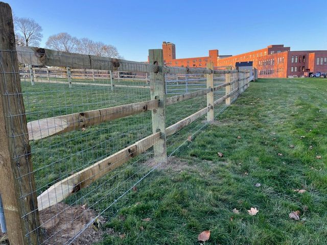 Wooden fence beside a grassy field with red brick buildings under a blue sky