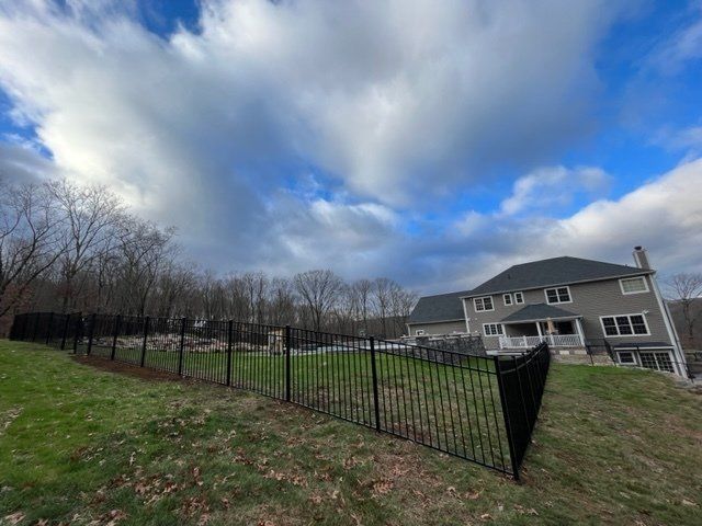 Fenced backyard beside a gray house under a cloudy blue sky, with bare trees and grassy lawn