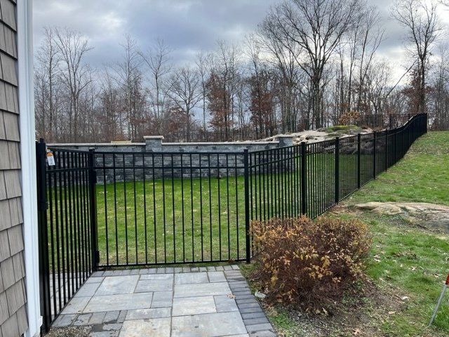 Black metal fence and gate beside a paved patio and grassy yard with bare trees in the background