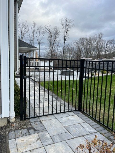 Black metal gate opening to a paved path and green lawn in a residential yard under a cloudy sky