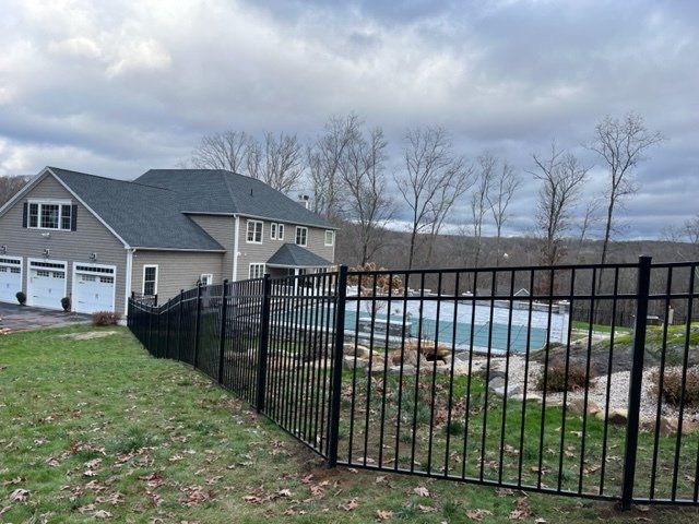 Black metal fence around a backyard pool beside a gray house under a cloudy sky