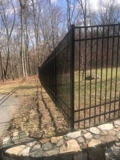 Black metal fence along a stone border beside a wooded path in a leafless park.
