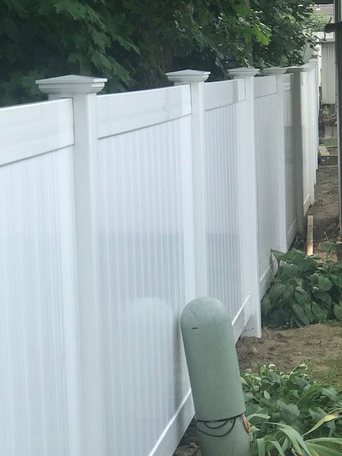 White vinyl privacy fence beside a narrow dirt path, with green foliage and a green post in front.