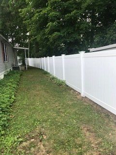 Narrow grassy side yard beside a house with a long white fence and trees overhead