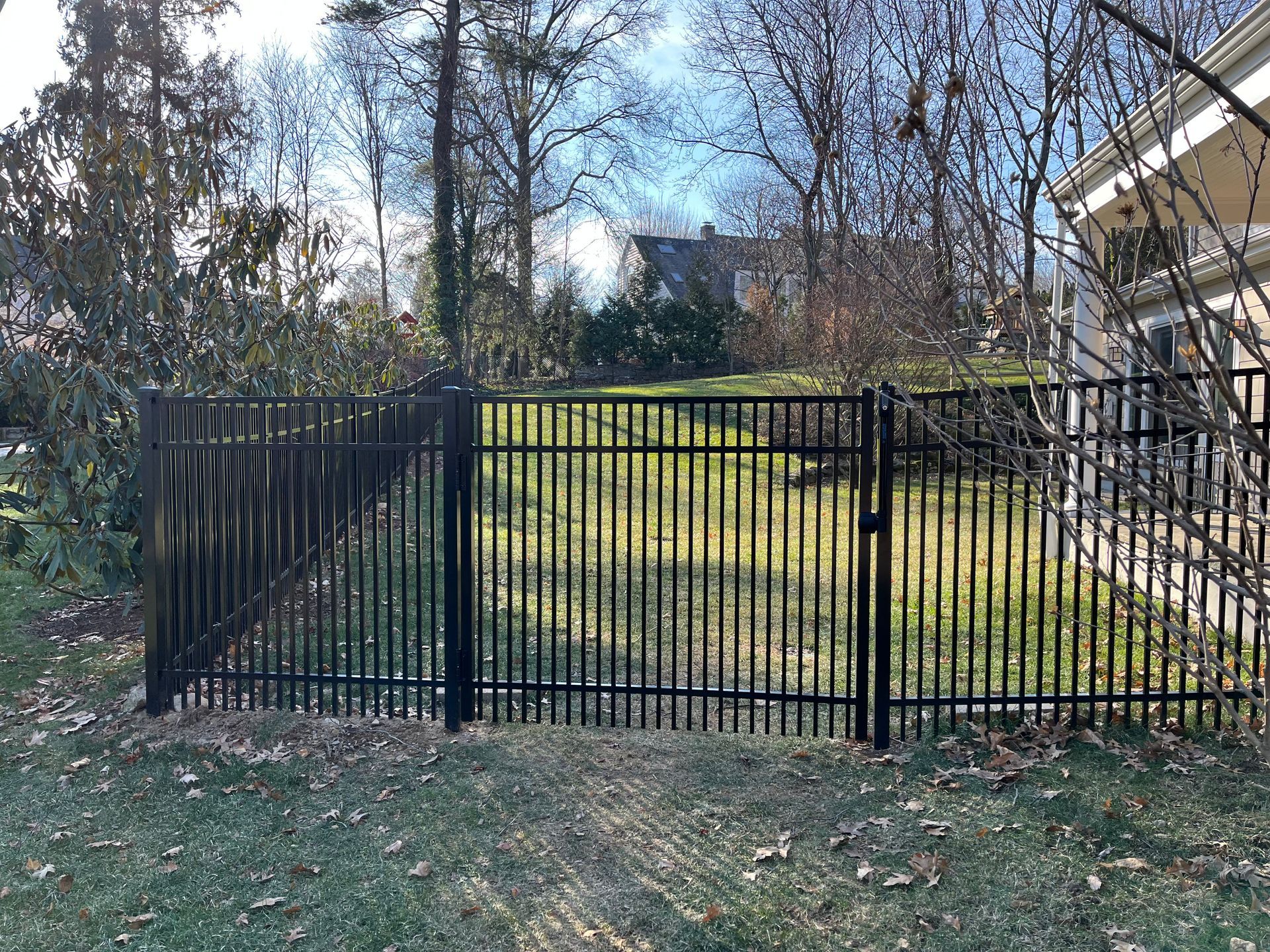 Black metal fence across a grassy yard with trees and a house at the right edge