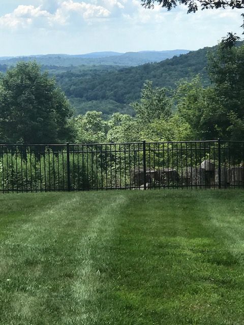 Lush green lawn leading to a black fence, with rolling forested hills under a cloudy sky