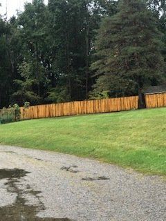 Gravel driveway beside a grassy yard and orange wooden fence, with tall trees in the background.