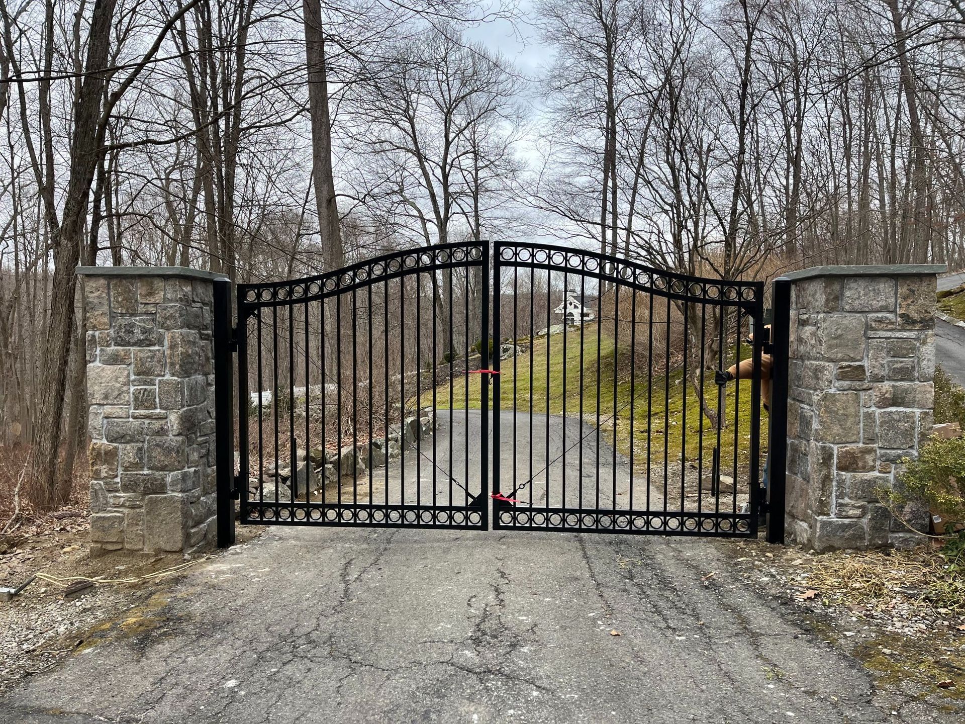 Black wrought-iron gate between stone pillars across a gravel driveway in a leafless wooded area