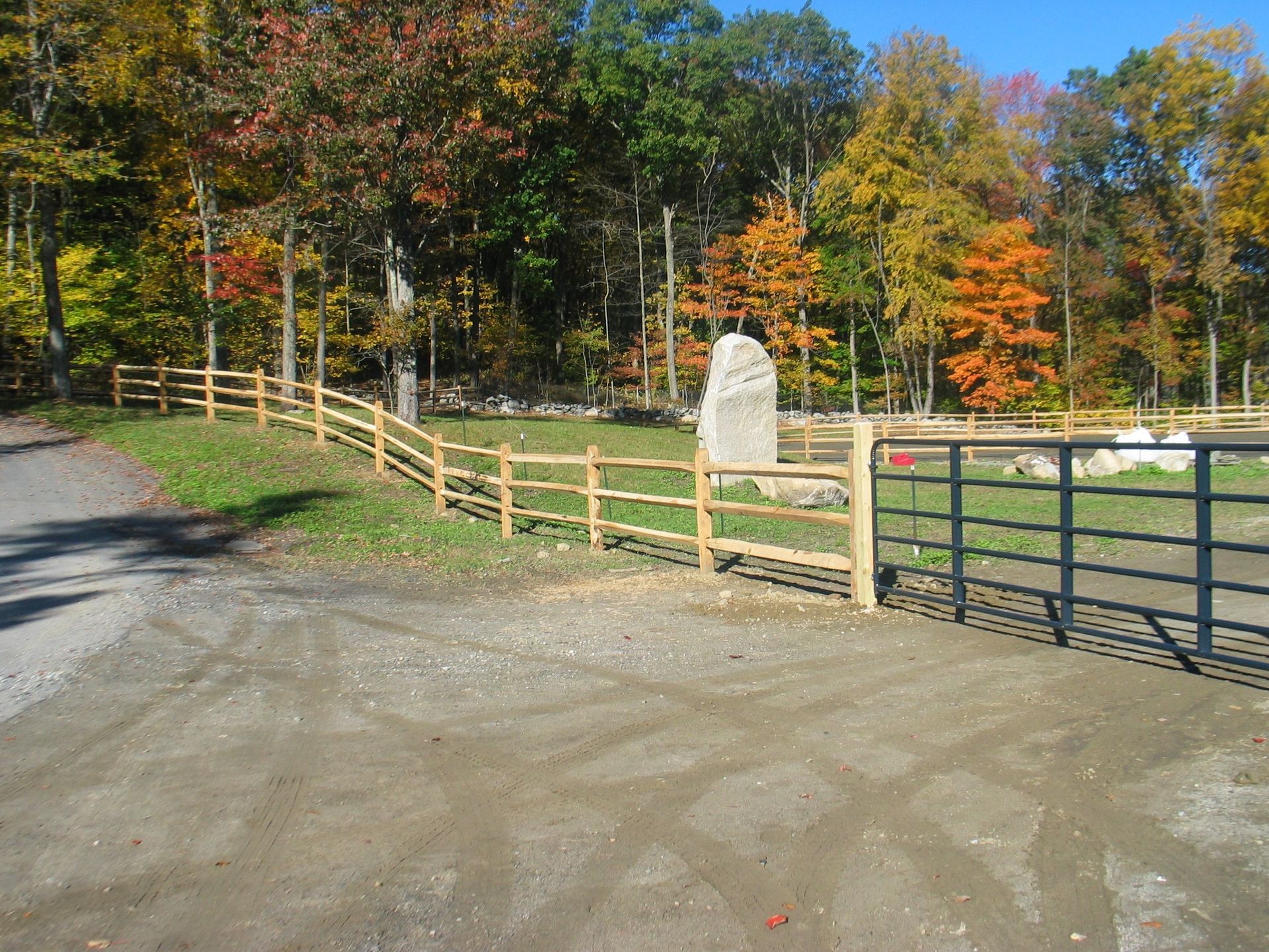 Gravel driveway beside a wooden fence and gate, with autumn trees and a stone marker in the background