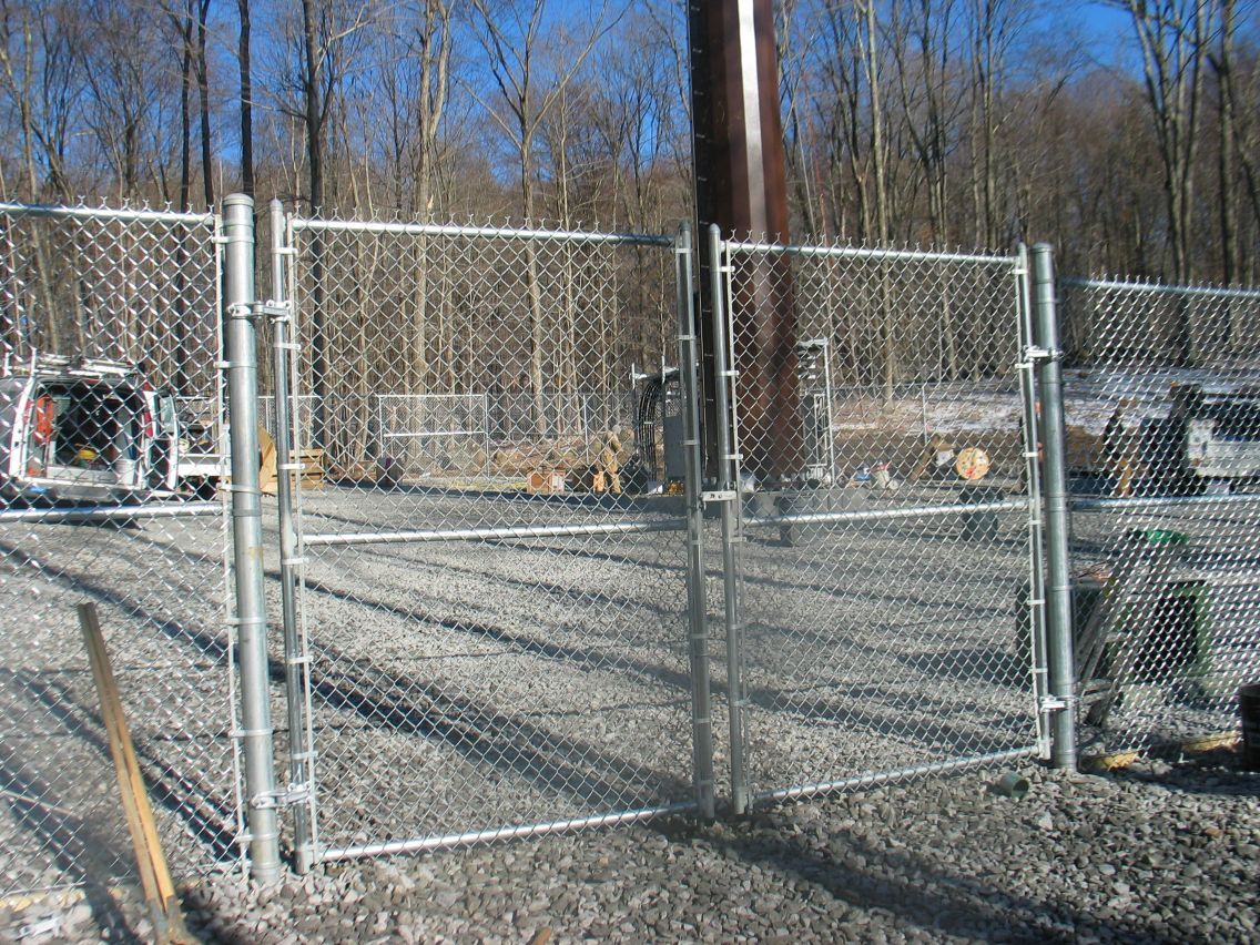 Chain-link gate and fencing at a gravel construction site with bare trees in the background