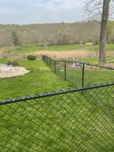 Black chain-link fence beside a grassy yard overlooking a pond and wooded hillside