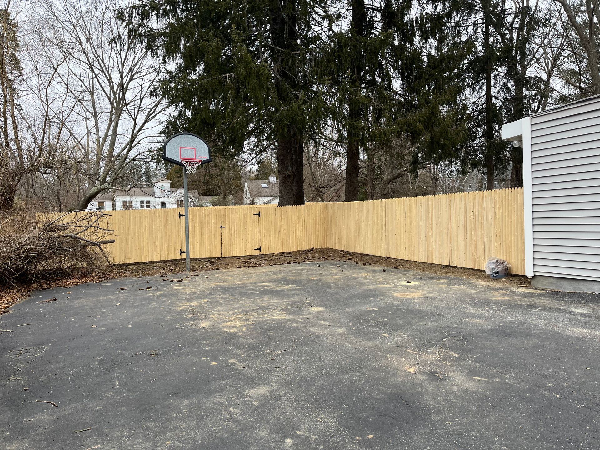 Empty paved backyard with a wooden fence, basketball hoop, trees, and a white shed