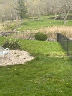 Backyard lawn with patio chair, gravel bed, black fence, and a pond bordered by trees.