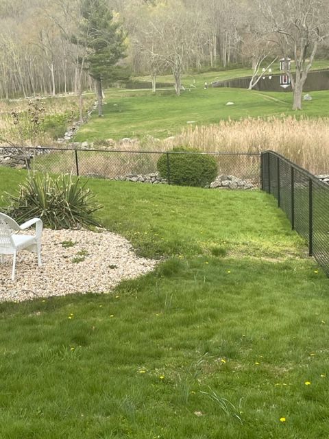 Grassy backyard with a small pond, black fence, gravel patio, and trees in the background