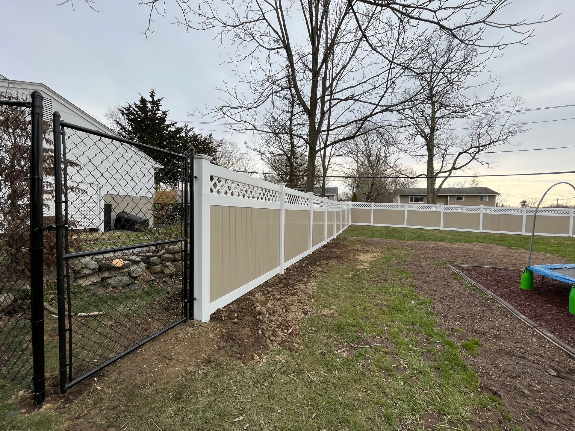 Chain-link and beige privacy fence along a grassy backyard under bare trees, near a house at dusk
