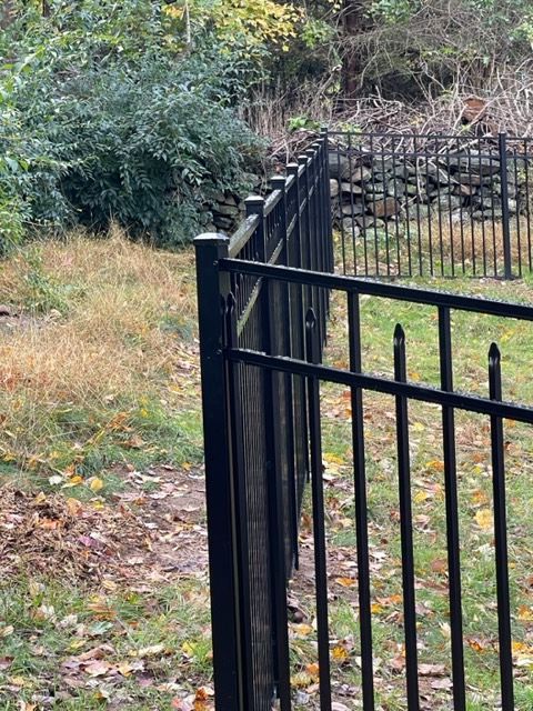 Black metal fence beside a grassy path with trees and a stone wall in the background