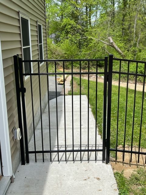 Black metal gate on a concrete patio beside a house, opening to a grassy yard and trees.