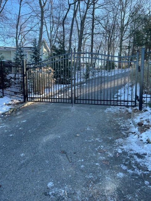 Snow-covered driveway leading to a black metal gate and fenced yard with bare trees.
