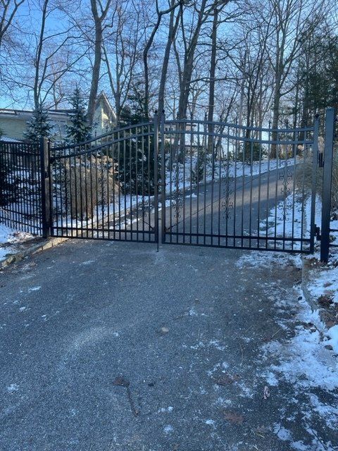 Snowy driveway with a black metal gate and leafless trees in the background