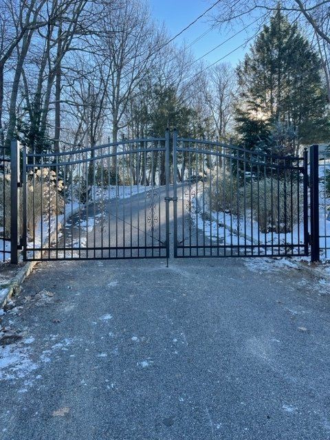 Black metal gate blocking a snowy driveway lined with leafless trees under a blue sky
