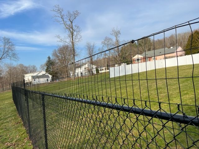 Chain-link fence beside grassy field with houses and trees under a blue sky