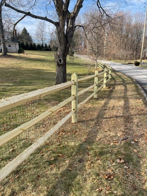 Wooden split-rail fence curving along a grassy roadside with leafless trees under a blue sky
