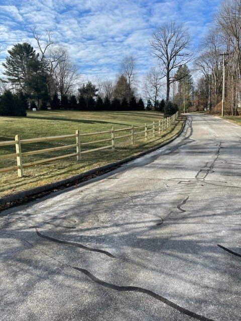 Curving paved road beside a wooden fence and grassy field under a blue, partly cloudy sky