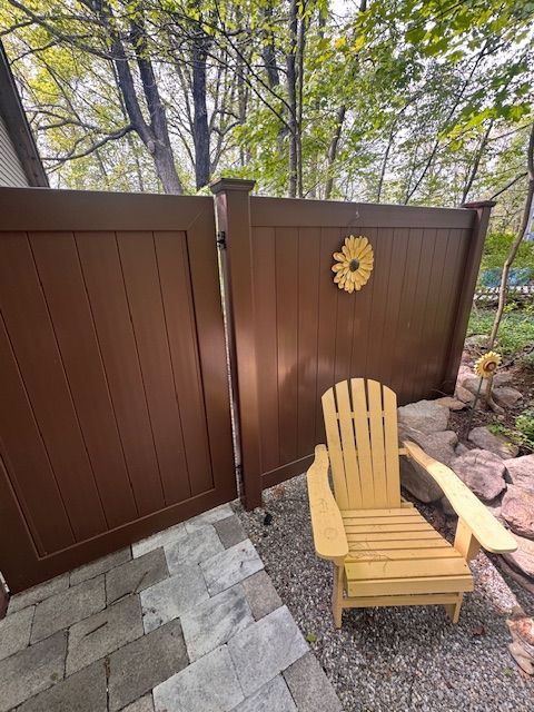 Wooden Adirondack chair on gravel beside a brown fence and gate, with stone pavers and trees beyond.