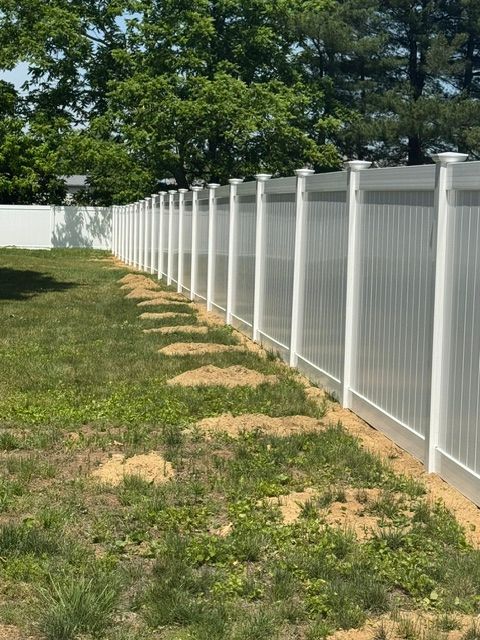 White vinyl fence lining a grassy backyard with trees and patchy dirt under a clear sky