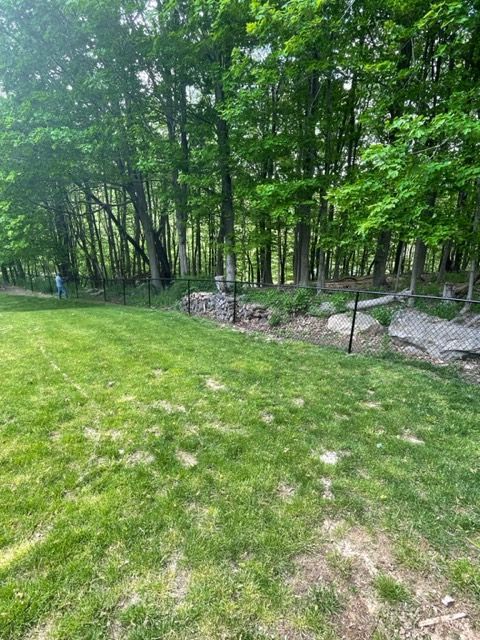 Grassy clearing beside a wooded trail with a small stone-lined stream and fence.