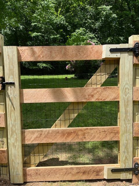 Wooden farm gate with black hinges and diagonal brace, set against green trees and grass.