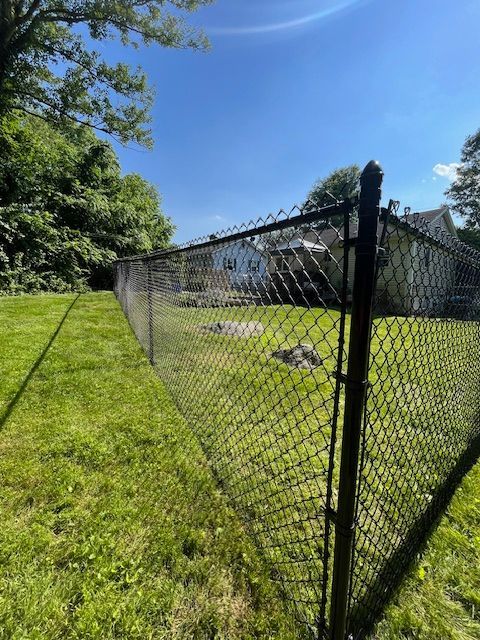 Chain-link fence along a sunny grassy yard beside a house under a blue sky.