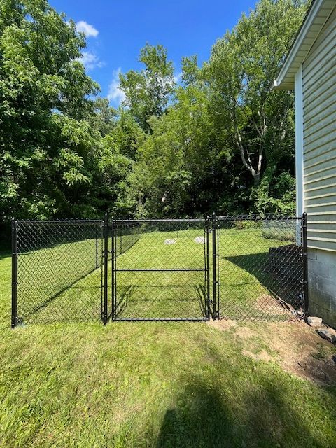 Black chain-link gate in a grassy backyard beside a white house, with trees and blue sky beyond.