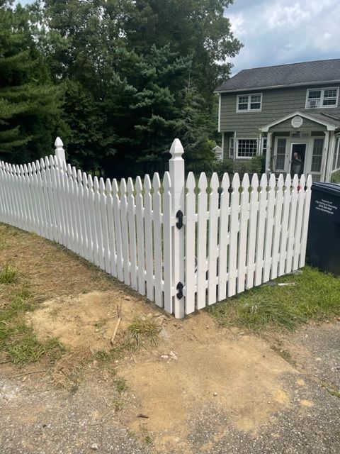 White picket fence with gate around a suburban house and yard, beside a dirt driveway