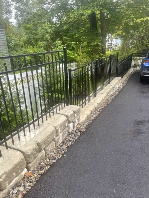 Black metal fence along a stone wall beside a wet roadside with trees; parked car at right