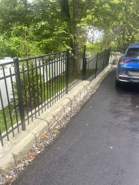 Parking lot beside a black metal fence and stone curb, with trees along the left and a blue car on the right.