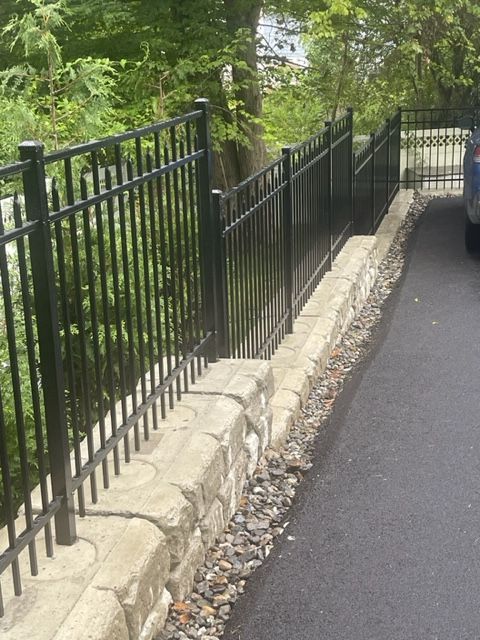 Black metal fence along a stone retaining wall beside a paved path in a leafy outdoor area