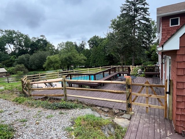 Wooden outdoor swimming pool beside a red house, with deck, fence, and trees under cloudy sky